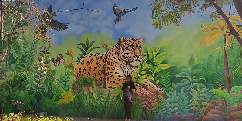 A woman stands before a detailed mural depicting a lush jungle, filled with vibrant plants and wildlife, creating a lively backdrop.